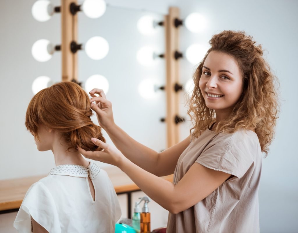 Female hairdresser smiling making hairstyle to beautiful redhead girl in beauty salon. Copy space.